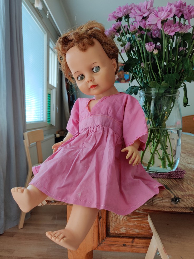 Ideal Doll nukke 1960-luvulta. Nukke istuu pöydällä. Nuken vieressä on kukkakimppu.